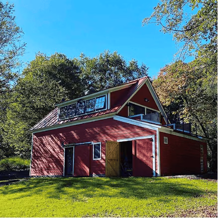 Red barn-style home with loft windows, open doors, and lush green lawn beneath a clear blue sky and trees.