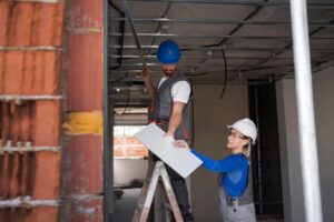 Two construction workers wearing safety helmets reviewing building plans at a construction site.