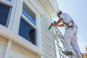 Painter cleaning or preparing a house exterior while standing on a ladder beside a window.